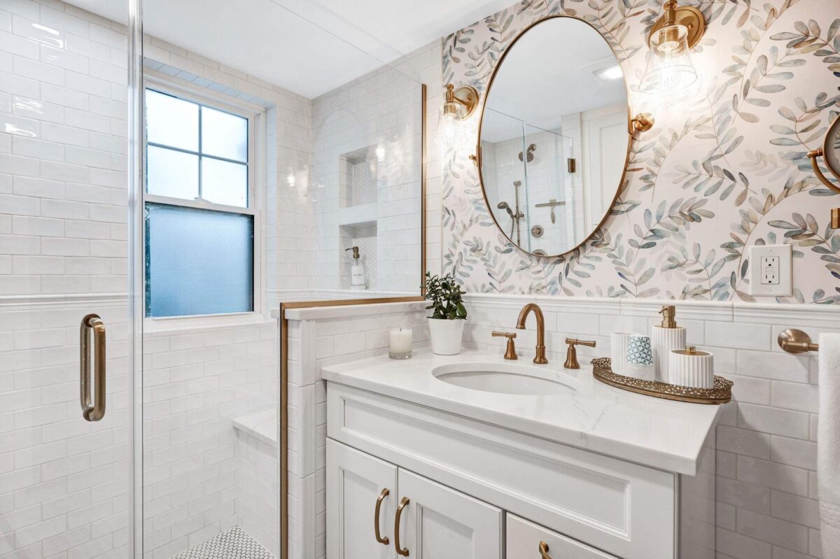 Bathroom with a white vanity, oval undermount sink, brass faucet and a round brass-framed mirror against leaf-pattern wallpaper