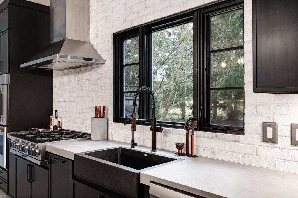 Black farmhouse sink in a white brick kitchen with black windows and a range hood