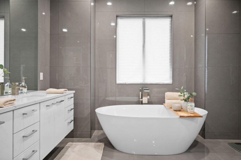 Freestanding white soaking tub in a modern bathroom with gray tile and a white vanity